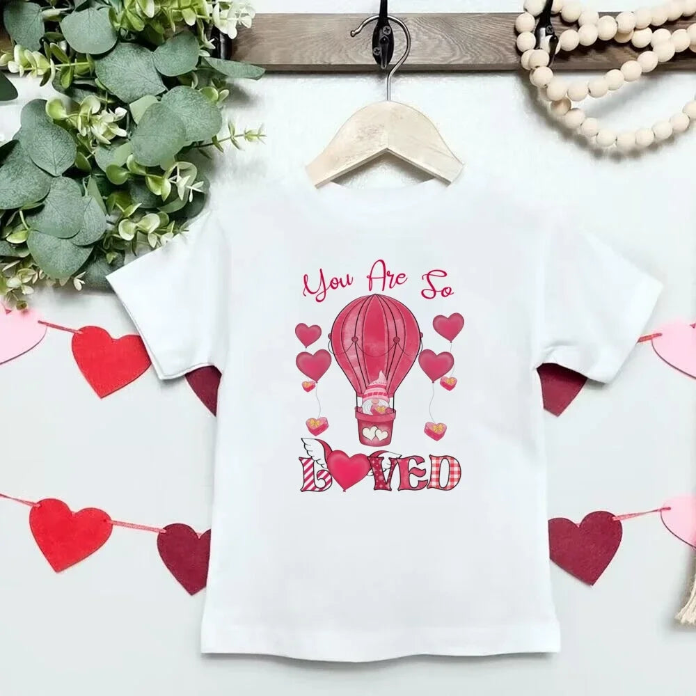 White t-shirt with pink hot air balloon and heart design on a hanger with decorative elements.