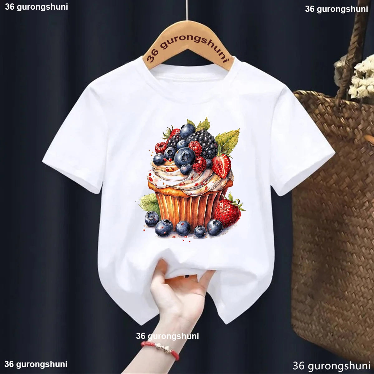 White t-shirt with a colorful cupcake graphic held on a hanger against a dark background.
