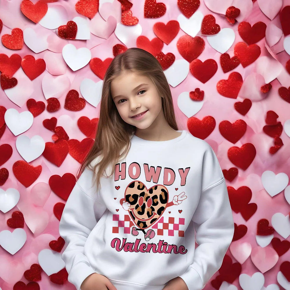 Child wearing a 'Howdy Valentine' sweatshirt in front of a heart-patterned background
