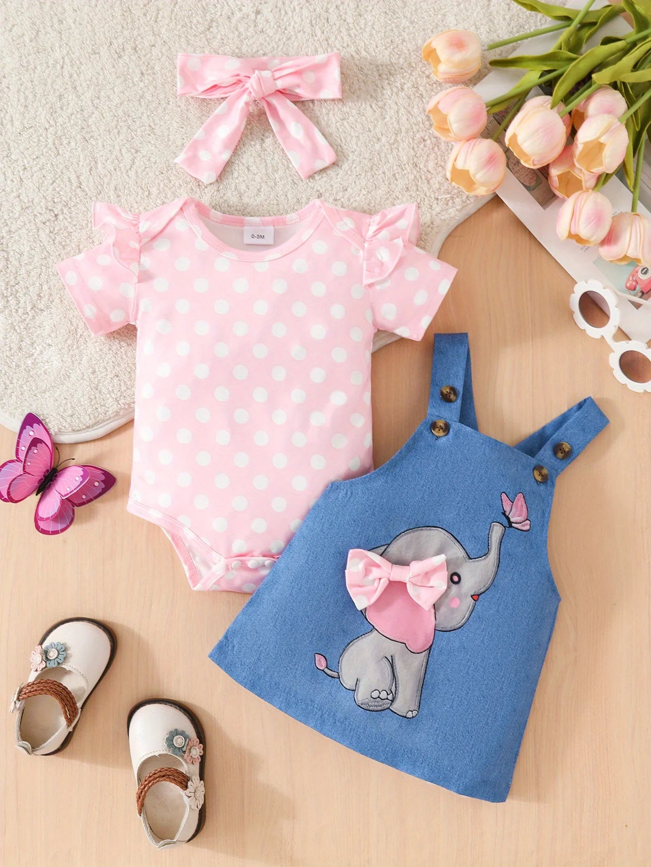 Baby outfit with pink polka dot onesie, blue dress with elephant design, and accessories on a light background.