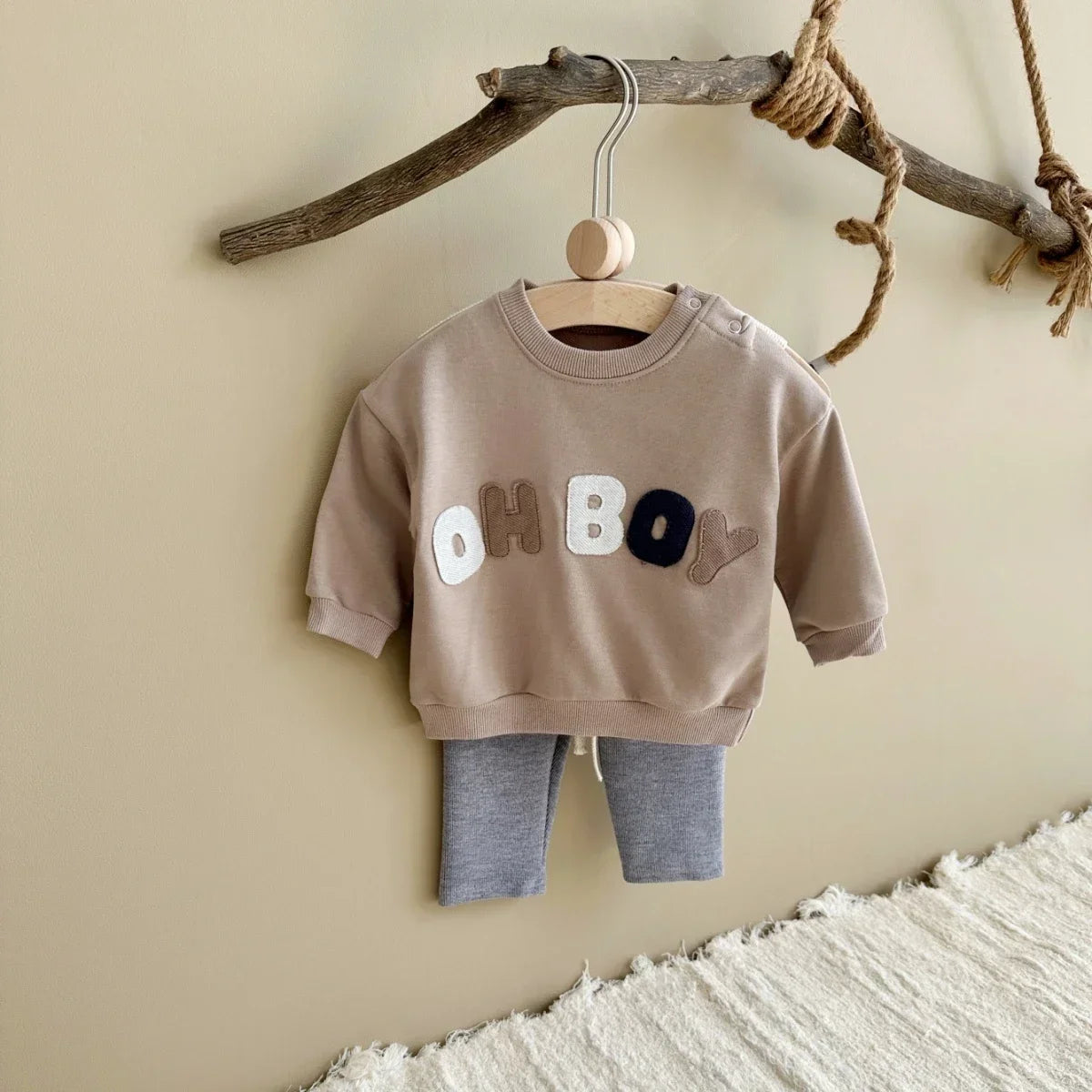 Children's brown sweatshirt with 'OH BOY' text and gray pants hanging on a branch against a beige wall.