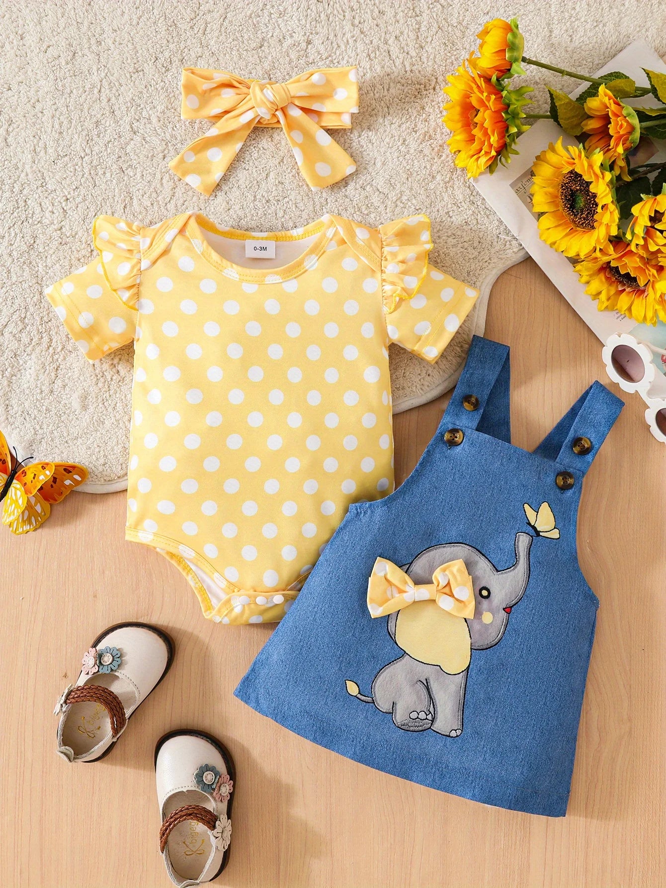 Yellow polka dot baby romper, blue denim dress with elephant design, and yellow headband on a wooden surface with flowers.