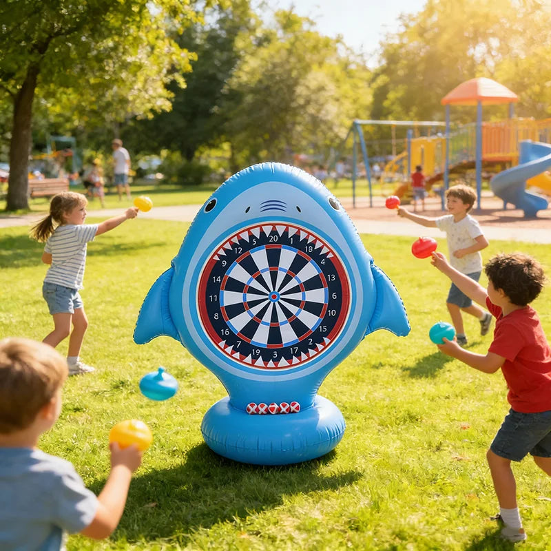 Children playing with a large inflatable shark dartboard on a sunny day at a park.
