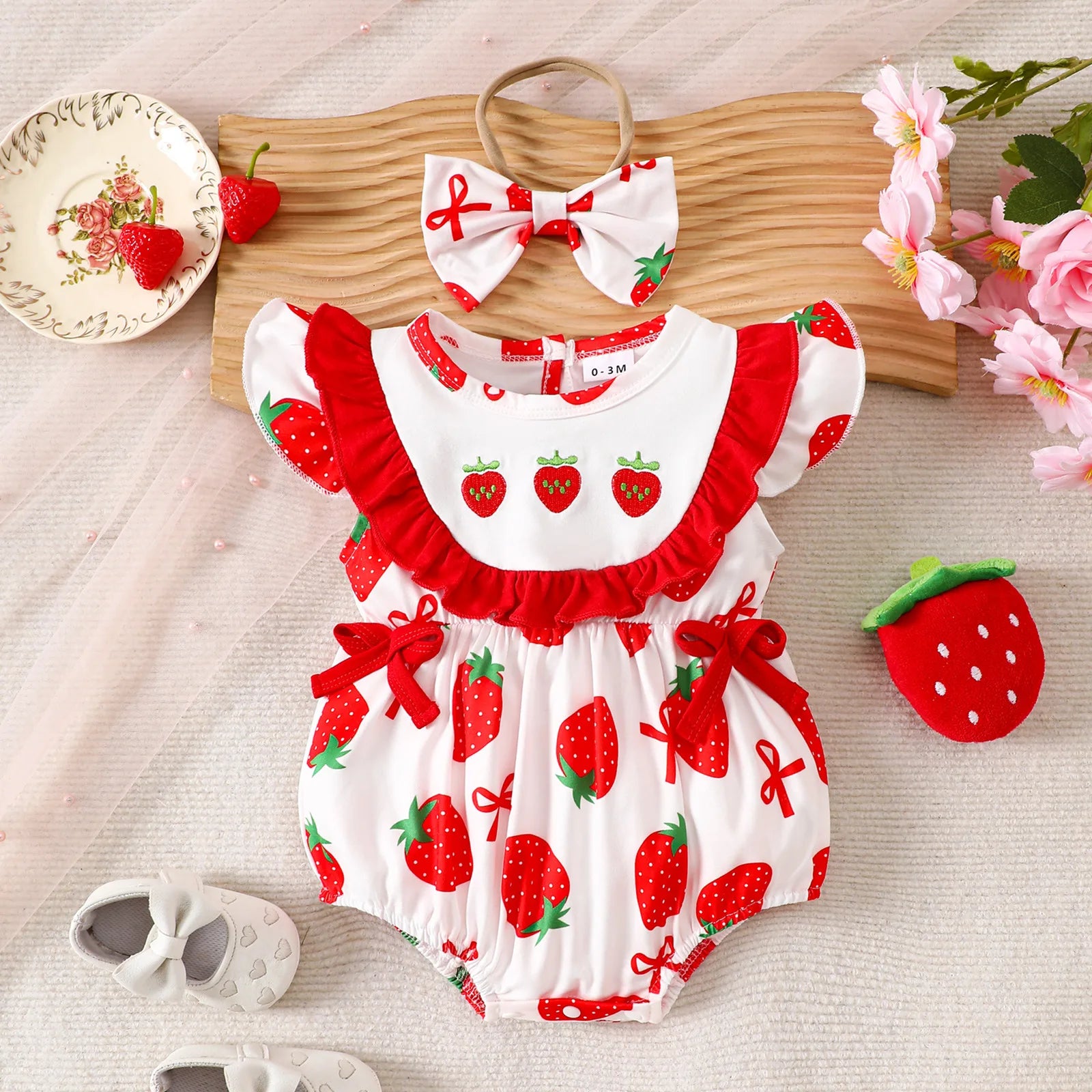 Baby romper with strawberry pattern, headband, and shoes on a wooden board with flowers.