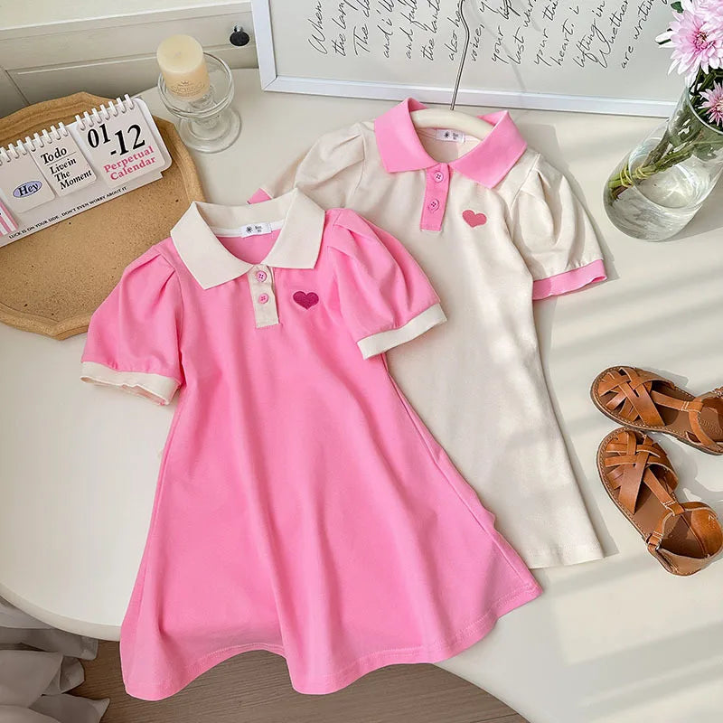 Two children's dresses, one pink and one white with pink collar, on a table.