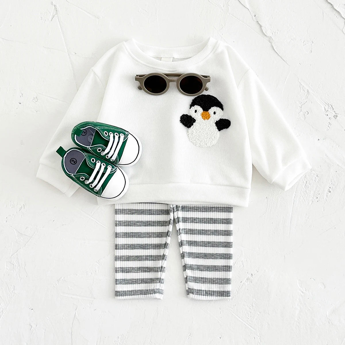 Children's outfit with white shirt featuring a penguin design, striped pants, and green shoes on a light gray background.